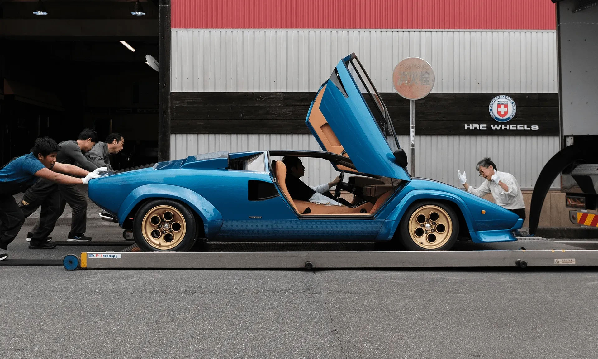 A blue Lamborghini Countach being loaded onto a transport truck outside a garage
